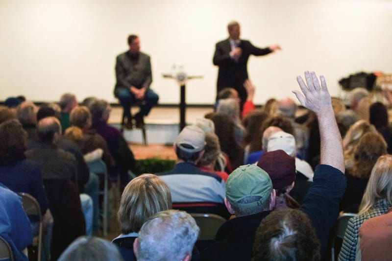 A person in a seated audience raises a hand to ask a question.