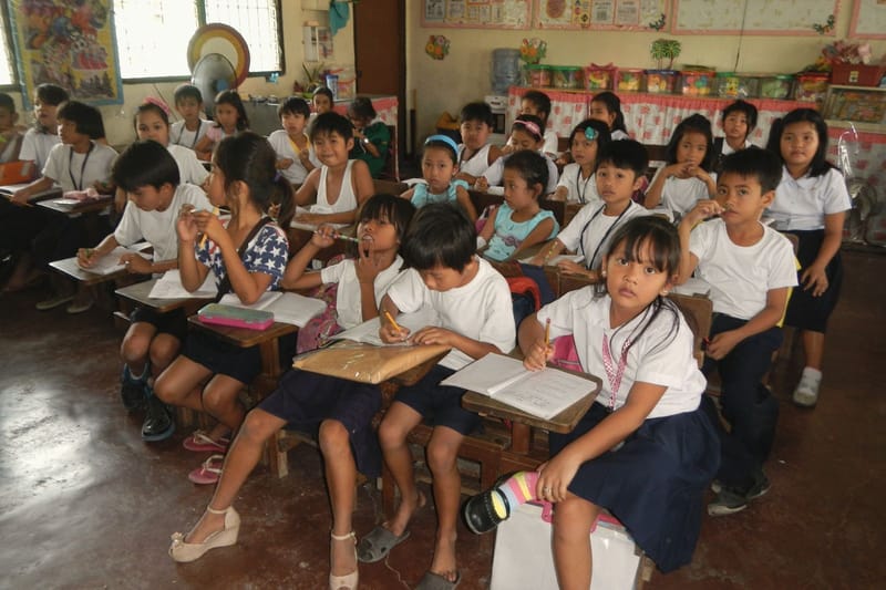 Elementary school children sit at their desks in a crowded classroom.